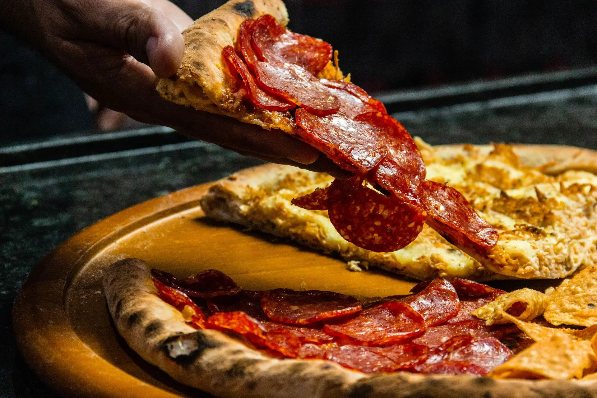Part de pizza au salame piccante soulevée d'une pizza entière posée sur une planche en bois, sortie du four.